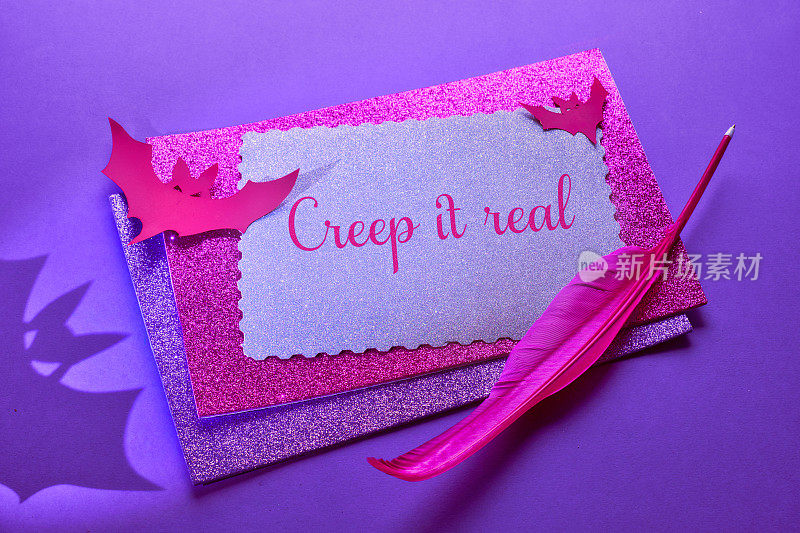 创意的紫色和粉色万圣节背景与堆叠的闪闪发光的卡片，纸工艺蝙蝠，粉色羽毛笔和长长的影子。背景上有蝙蝠的影子，文字“Creep it real”。